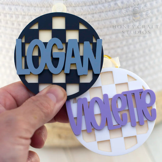 Two personalized checkered name tags, one black with slate blue text and one white with purple text, perfect for backpacks or school bags.