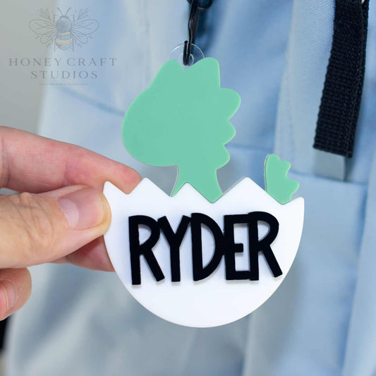 Green baby dinosaur name tag with bold black custom name Ryder in layered acrylic, attached to a backpack.
