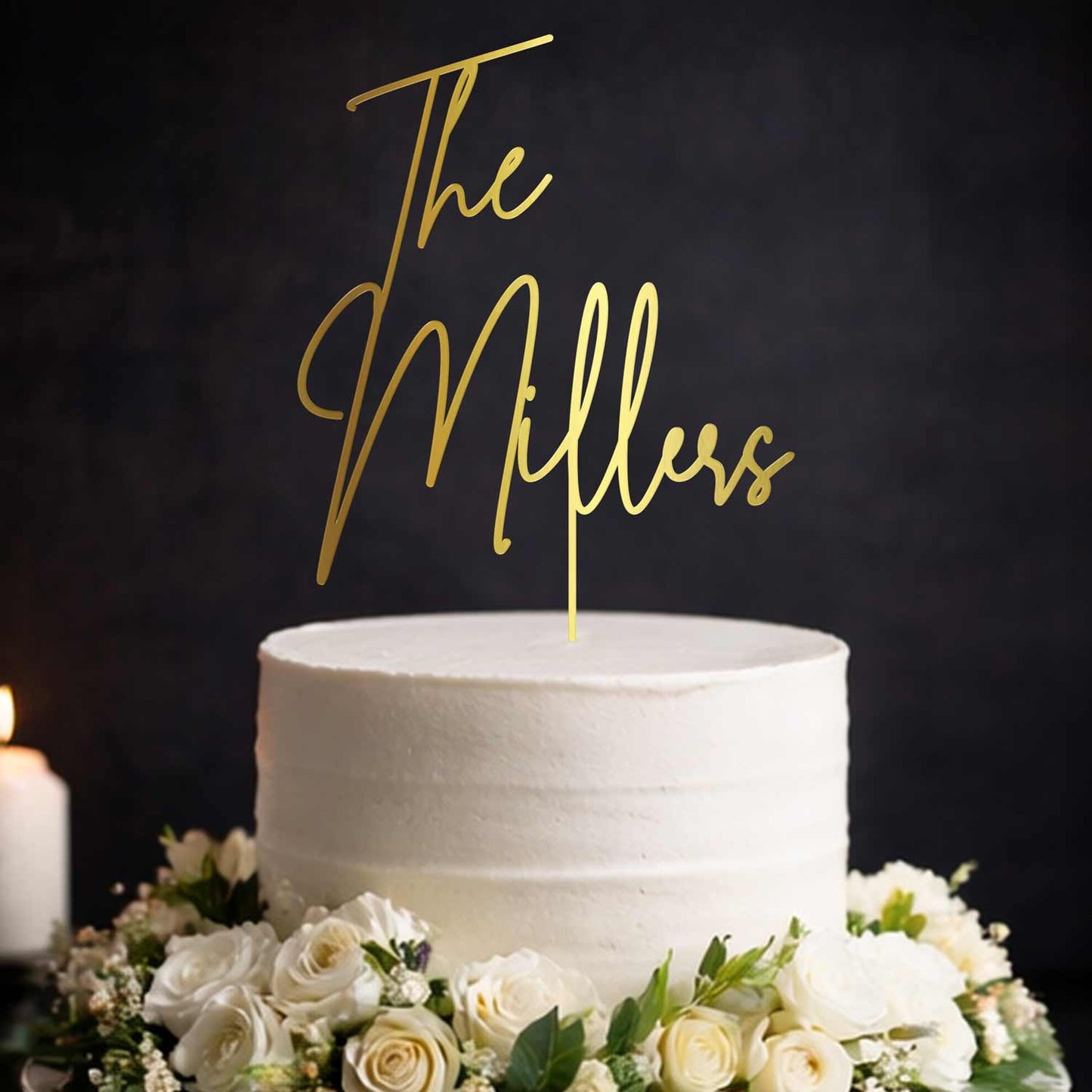 Gold mirror acrylic wedding cake topper with script text “The Millers” on elegant white wedding cake with florals. Made by Honey Craft Studios.