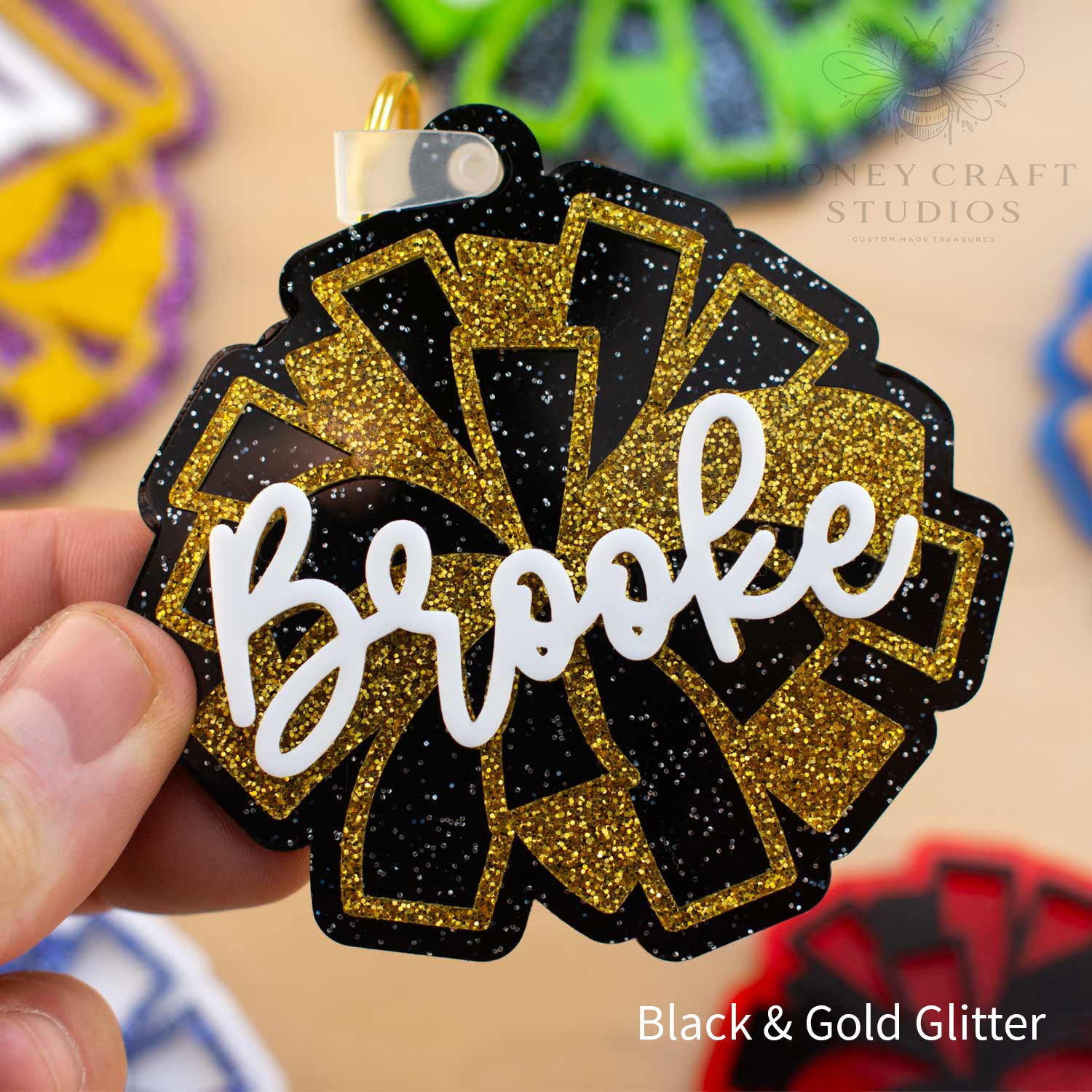 Personalized cheer pom-pom name tag keychain in black glitter and gold glitter acrylic, featuring the custom name “Brooke” in layered white script lettering.
