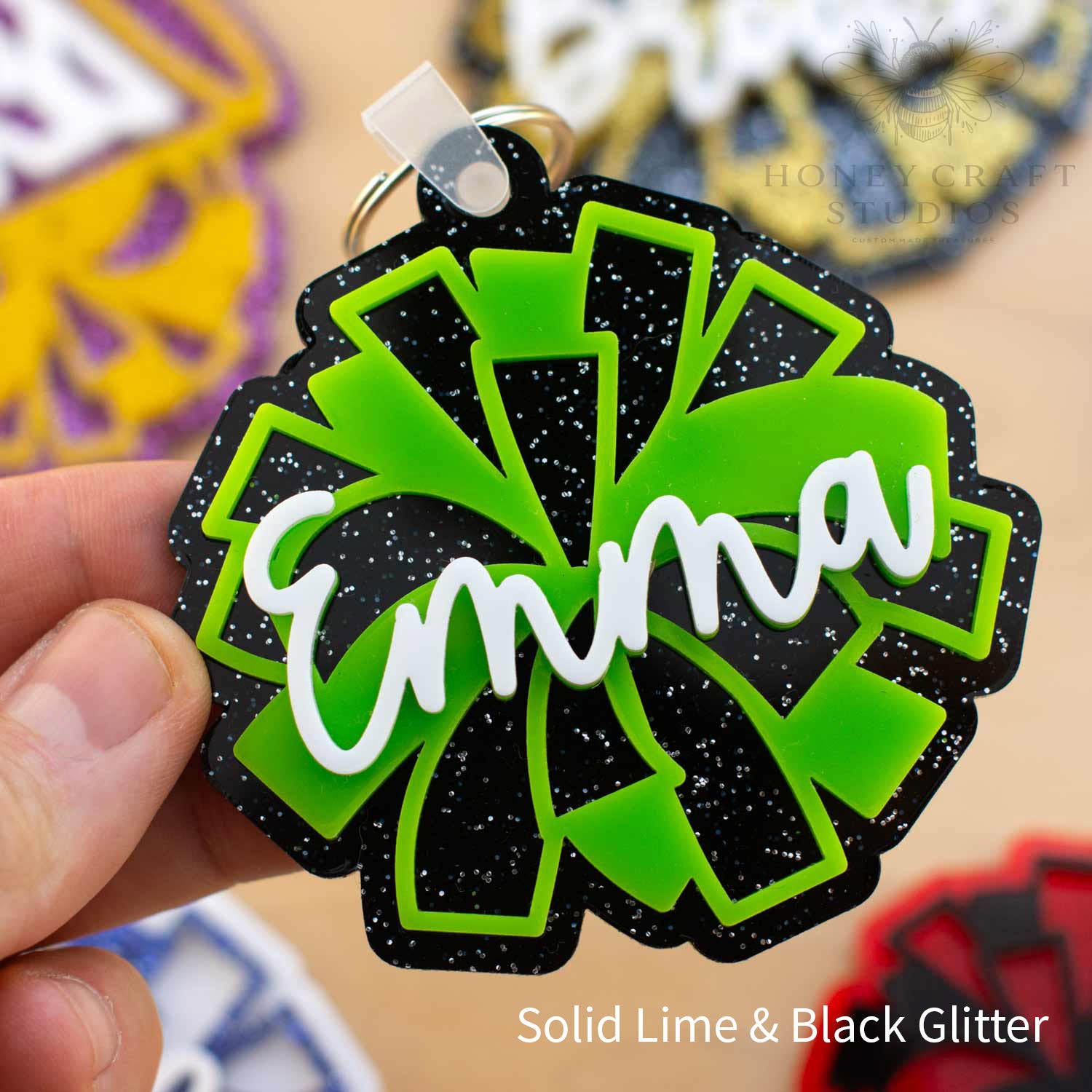 Custom cheer pom-pom backpack tag in black glitter acrylic with bright lime green accents, personalized with the name “Emma” in raised white script lettering.