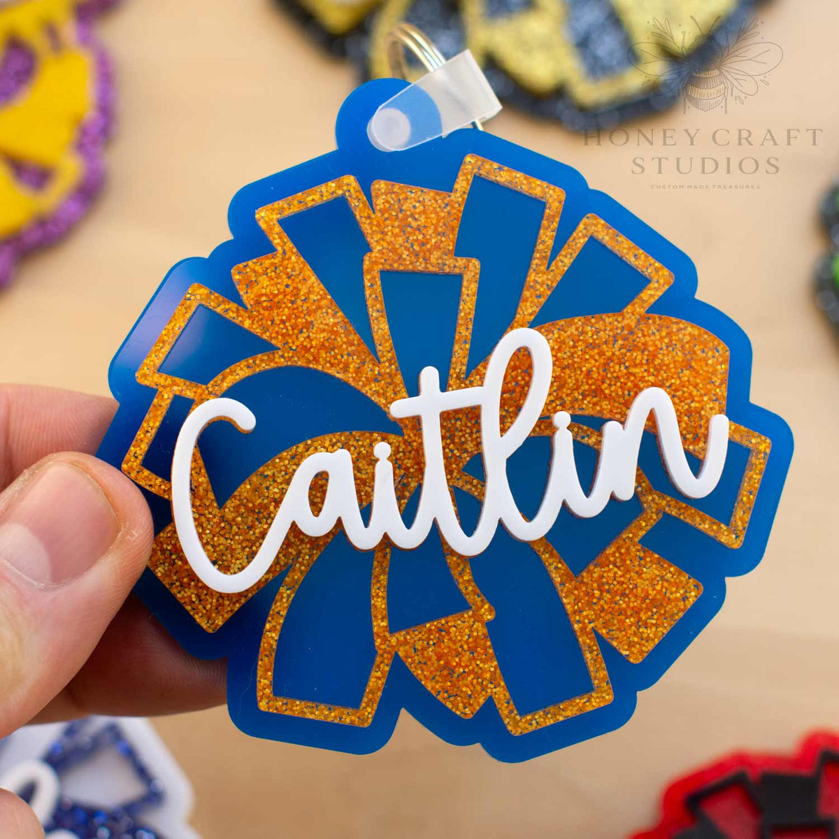 Personalized cheerleader name tag keychain in blue acrylic with orange glitter pom-pom details, customized with the name “Caitlin” in layered white script.