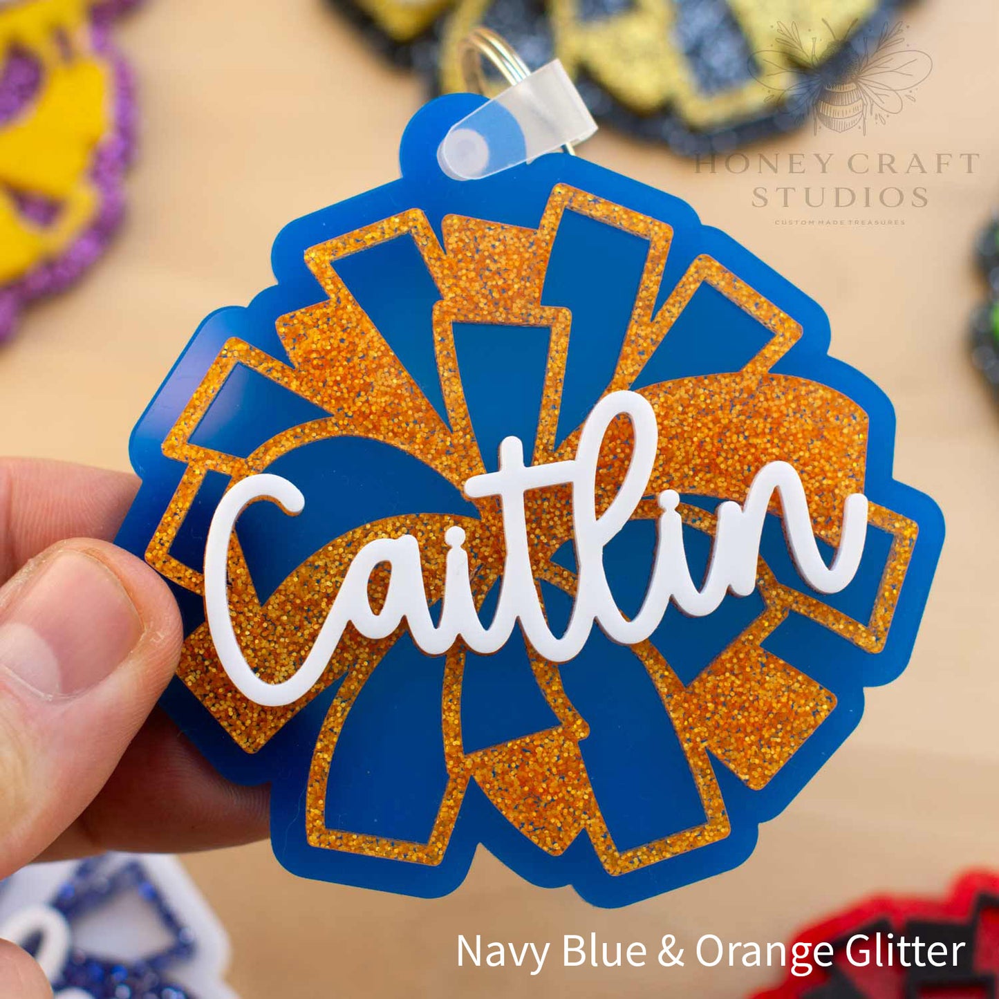 Personalized cheerleader name tag keychain in blue acrylic with orange glitter pom-pom details, customized with the name “Caitlin” in layered white script.