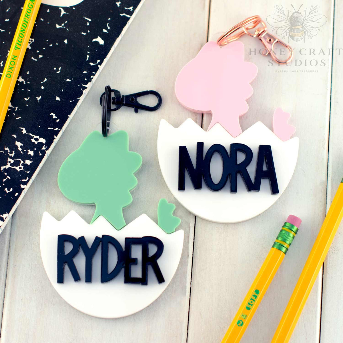 Personalized dinosaur name tags for kids shown in pink and green colors with names Ryder and Nora, ideal for backpacks or school bags.