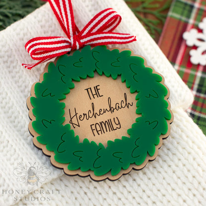 Personalized family Christmas wreath ornament with engraved wood center and green acrylic wreath, featuring red and white ribbon bow, handmade by Honey Craft Studios.