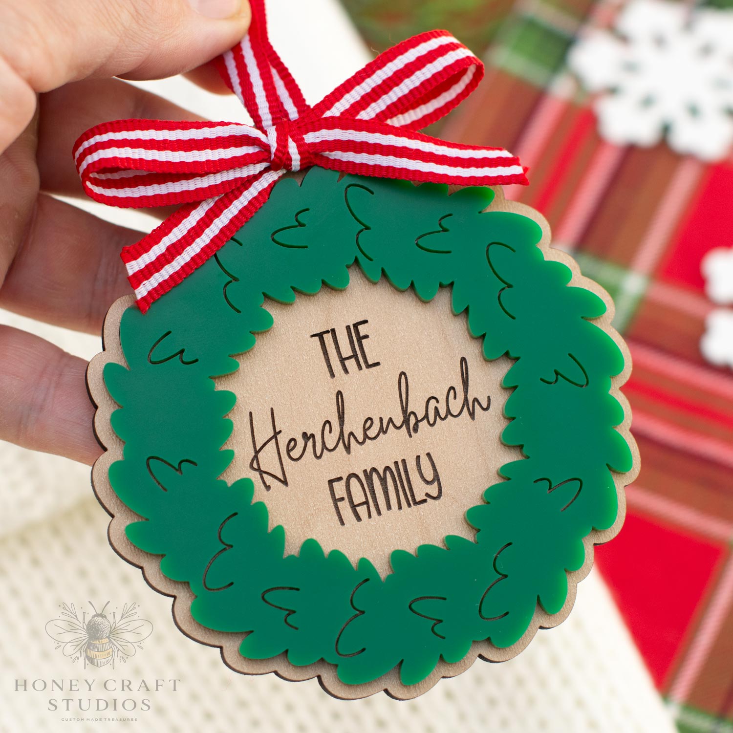 Hand holding a custom engraved family Christmas wreath ornament made from wood and green acrylic with red ribbon bow, personalized with family name by Honey Craft Studios.