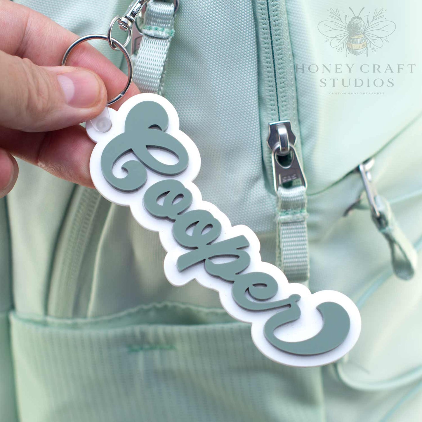 Personalized groovy-style name tag in fern green acrylic with white backing, attached to a keyring on a backpack bag.