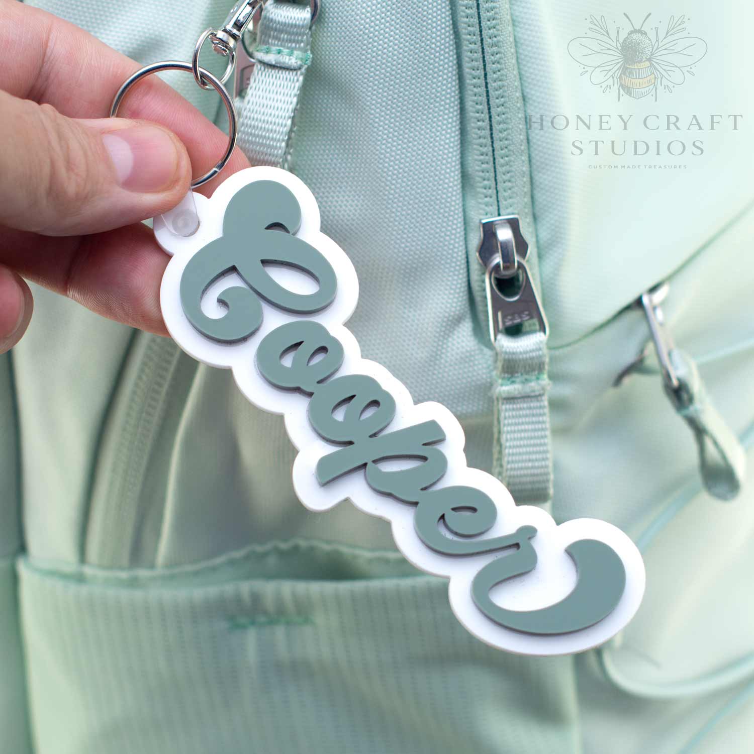 Personalized groovy-style name tag in fern green acrylic with white backing, attached to a keyring on a backpack bag.