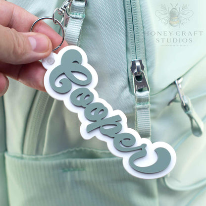 Personalized groovy-style name tag in fern green acrylic with white backing, attached to a keyring on a backpack bag.