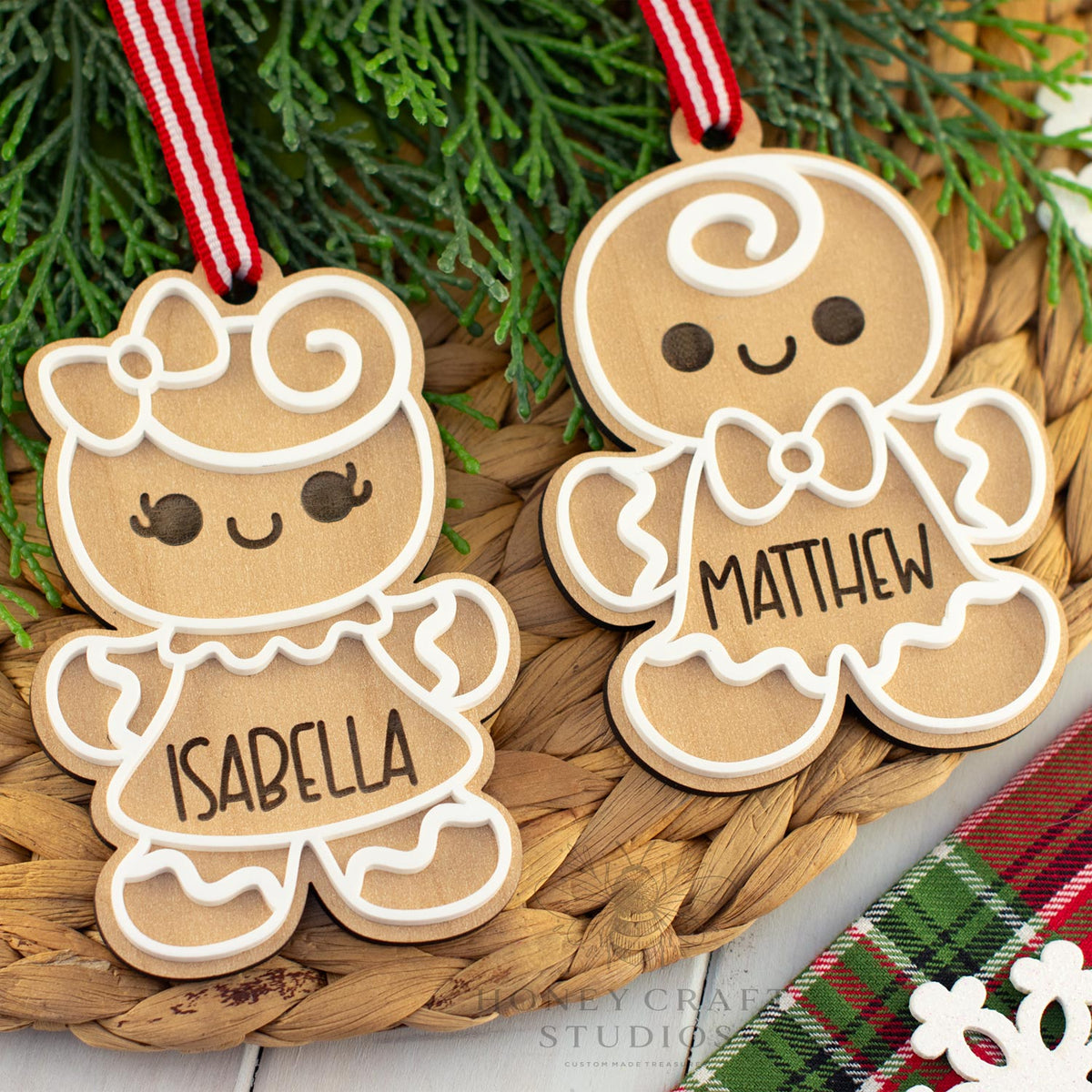 Two gingerbread-themed wooden ornaments with names 'Isabella' and 'Matthew' hanging on a woven mat with greenery in the background.