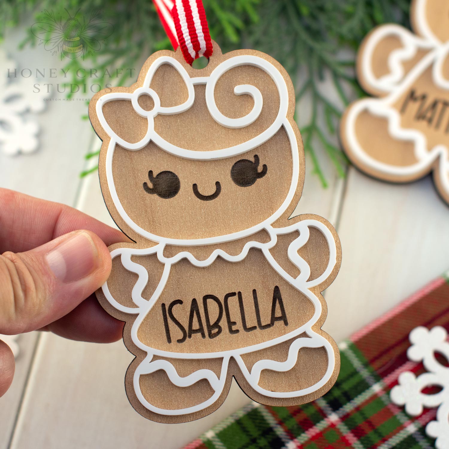 Hand holding a gingerbread girl ornament with 'Isabella' on it, Christmas tree in the background