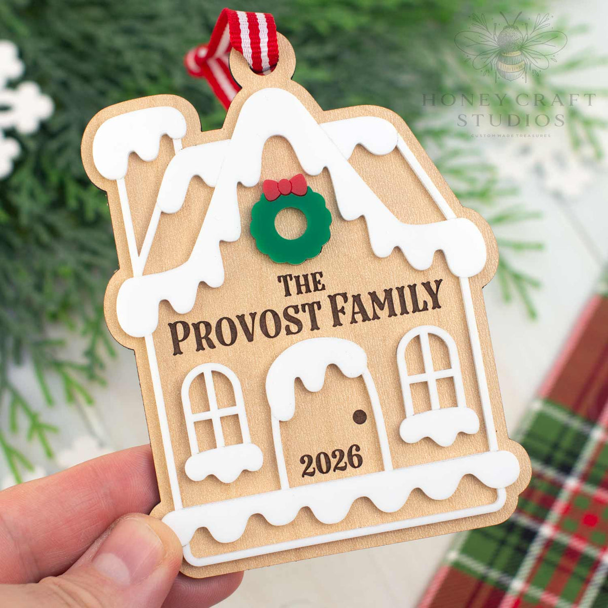 A hand holds a personalized wooden gingerbread house ornament with the text 'THE PROVOST FAMILY' and '2026', decorated with a green wreath and a red and white striped hanging ribbon. The background is a soft-focus holiday scene with green pine branches and a plaid blanket.