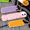 Personalized wooden Halloween name tags with names 'AGATHA', 'EMERSON', and 'MATEO' in pastel colors, decorated with black silhouettes of a witch, skull, and bat, displayed on a gray wood background.