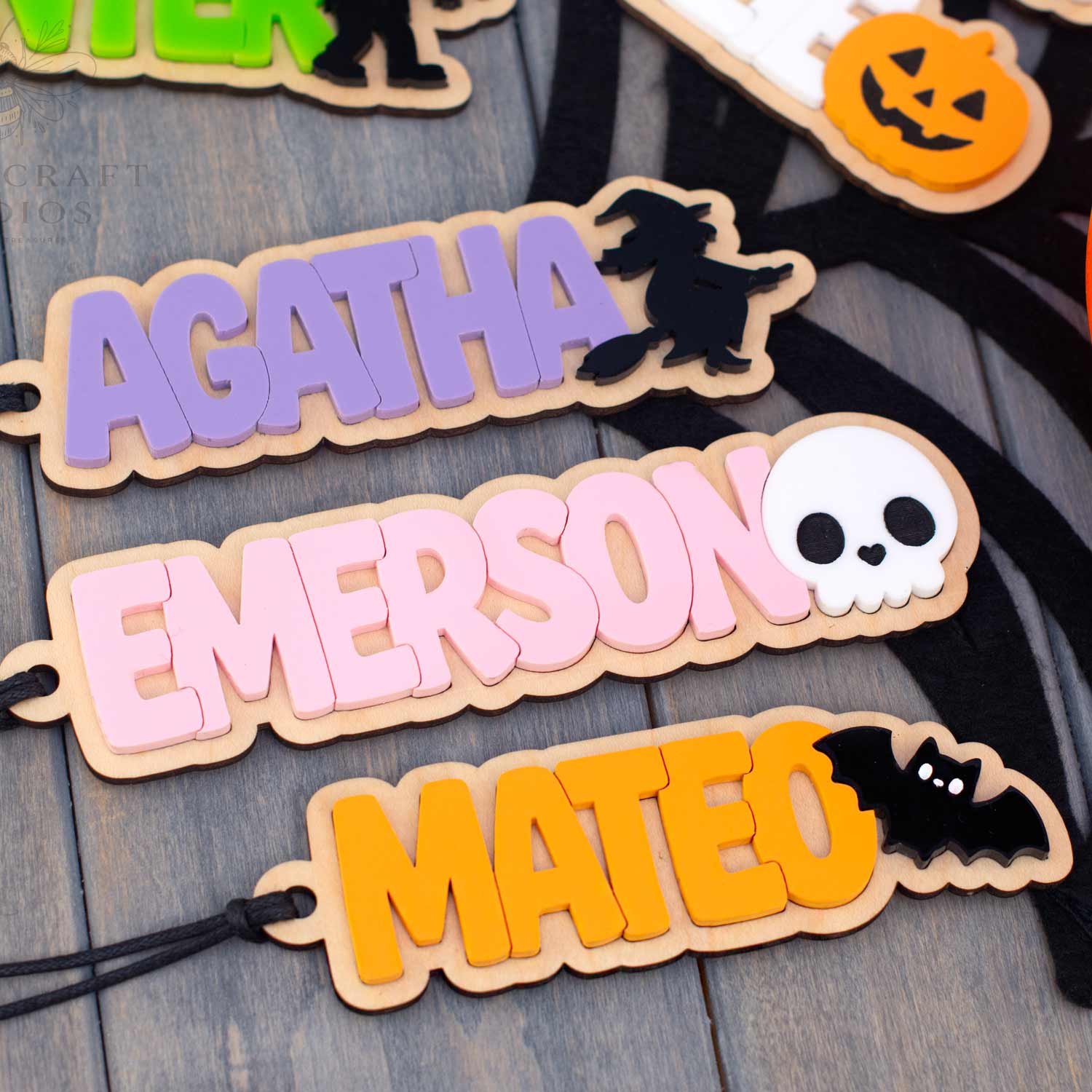 Personalized wooden Halloween name tags with names 'AGATHA', 'EMERSON', and 'MATEO' in pastel colors, decorated with black silhouettes of a witch, skull, and bat, displayed on a gray wood background.