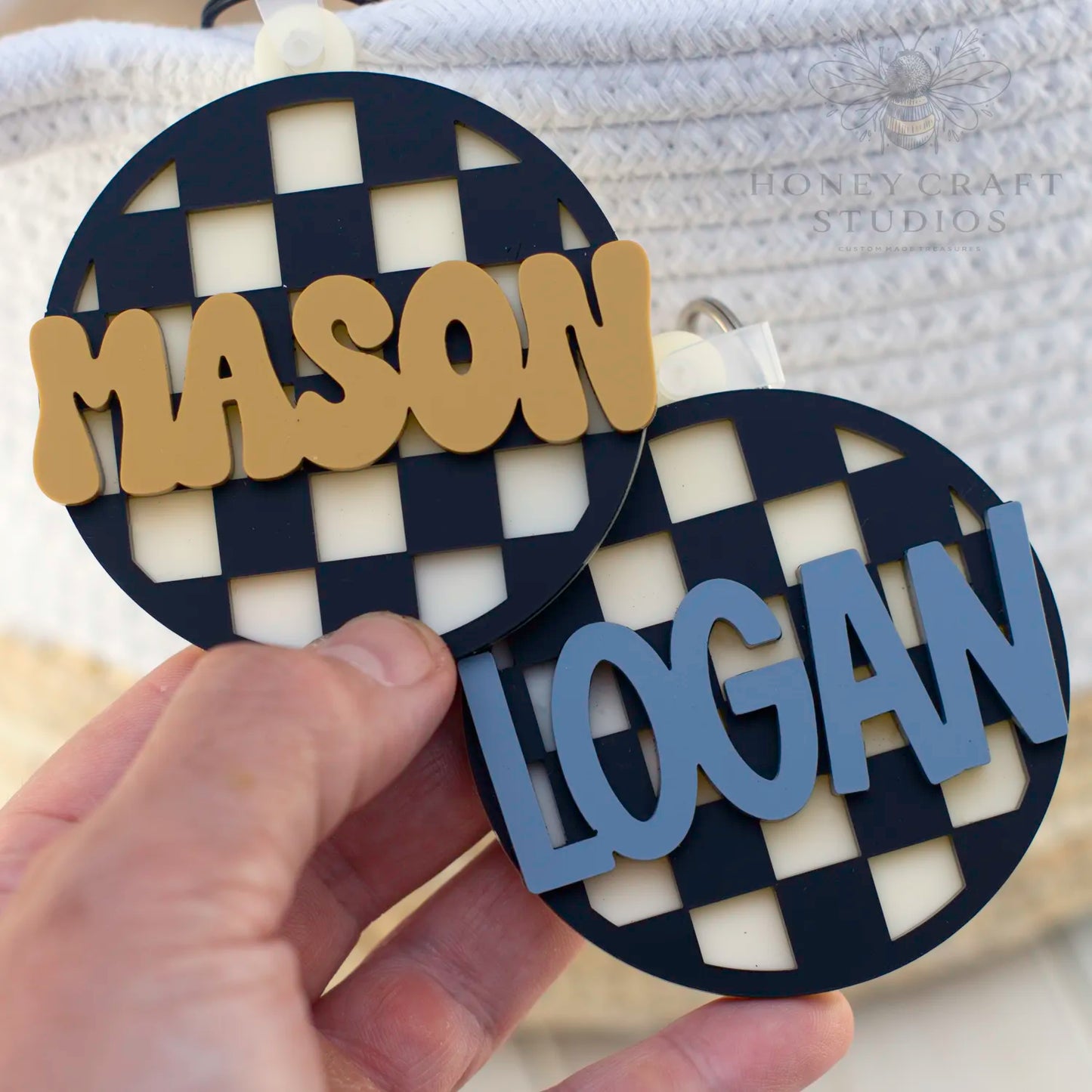 Side-by-side black checkered acrylic tags with personalized names in slate blue and mustard yellow text.