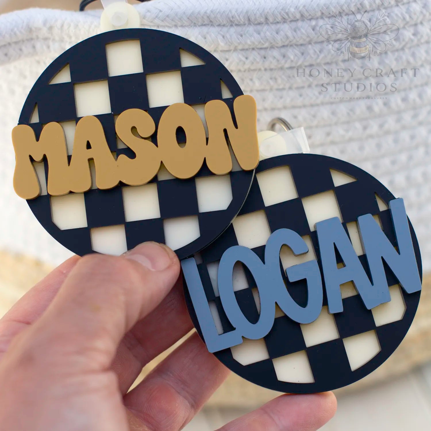 Side-by-side black checkered acrylic tags with personalized names in slate blue and mustard yellow text.