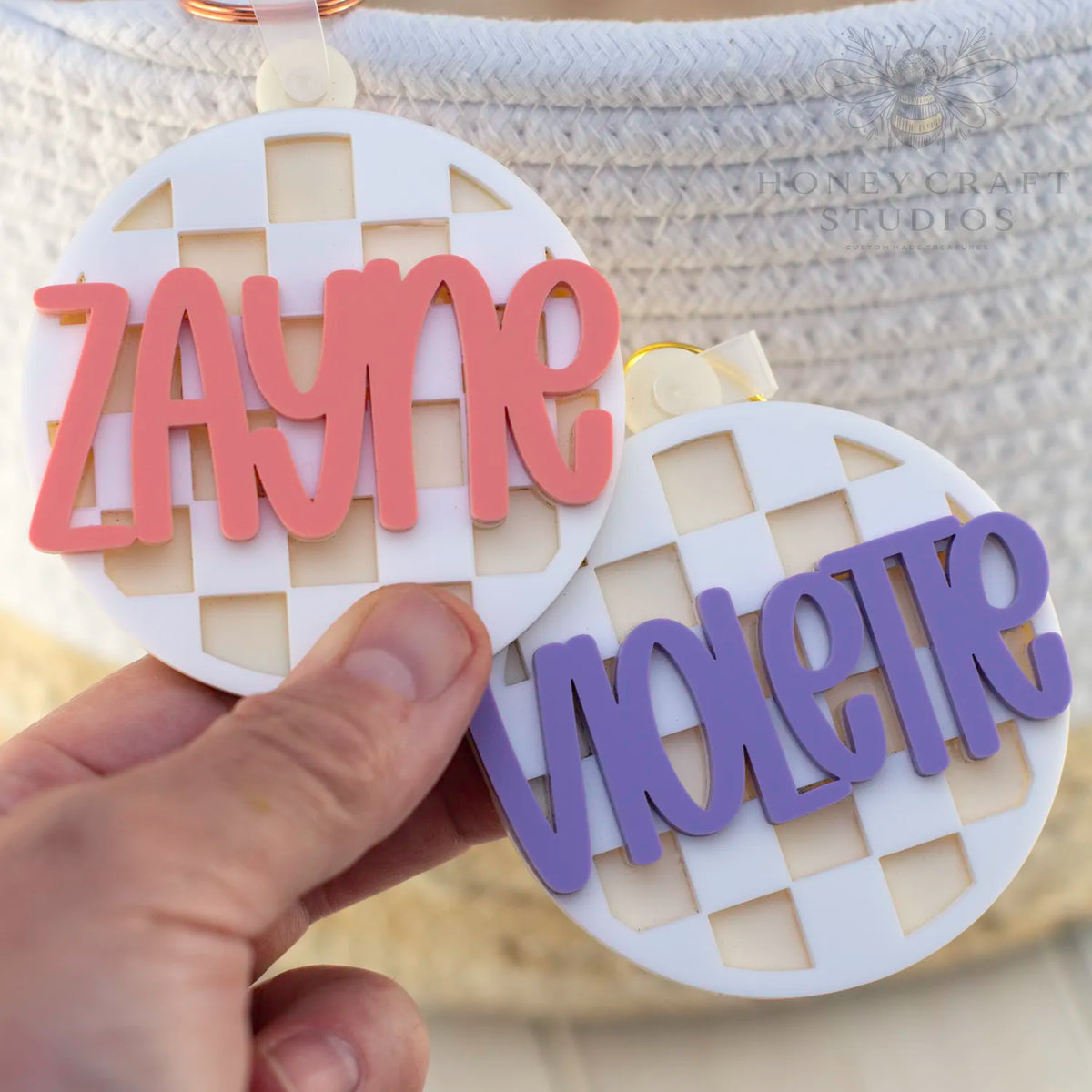 Two white checkered acrylic tags shown with custom names in pink and purple text, perfect for labeling kids’ gear.