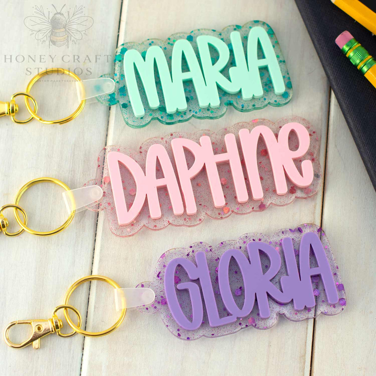 Three glitter acrylic name tags in teal, pink, and purple, labeled Maria, Daphne, and Gloria, with gold key rings on a white wood background.