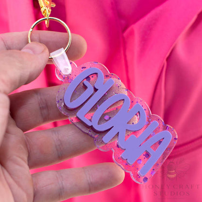 Purple Lilac glitter name tag labeled “Gloria” hanging from a backpack, showing layered acrylic detail.