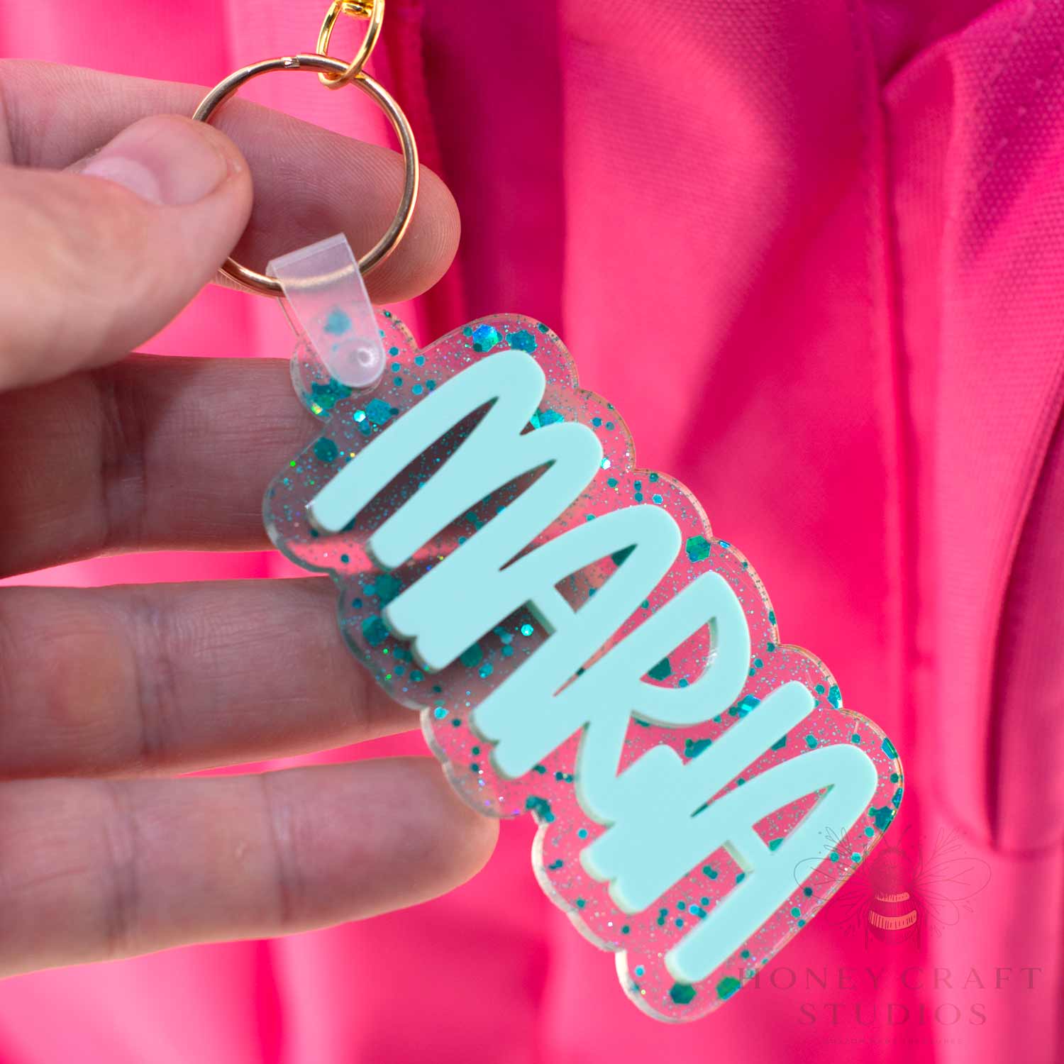 Custom teal glitter acrylic name tag reading “Maria” clipped to a school backpack.