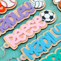 Personalized wood and acrylic sports name keychains with layered script names and engraved ball icons on teal wooden background