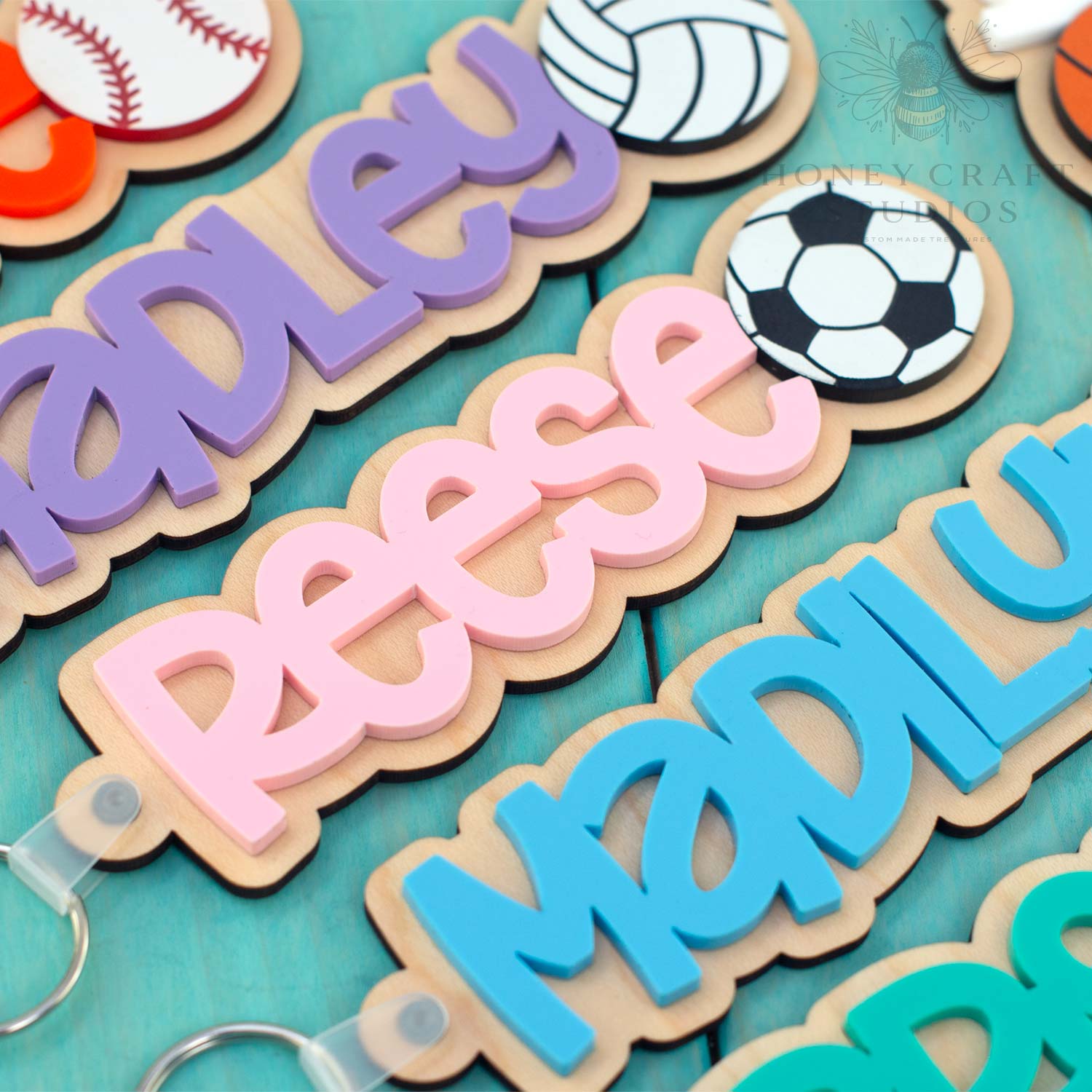 Personalized wood and acrylic sports name keychains with layered script names and engraved ball icons on teal wooden background
