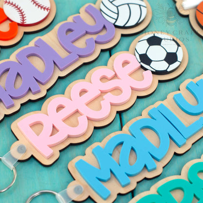 Personalized wood and acrylic sports name keychains with layered script names and engraved ball icons on teal wooden background