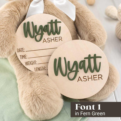 Two round personalized wooden baby keepsake tags with engraved text and lines, displayed on a plush teddy bear. The tags are light natural wood with dark green script text reading 'Wyatt ASHER'.