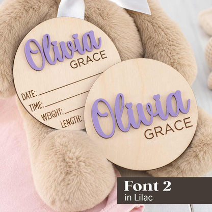 Two round wooden personalized tags with laser-cut text reading 'Olivia Grace' in lilac and brown, displayed on a plush beige teddy bear. One tag is filled in with date, time, weight, and length lines, and the other is blank.