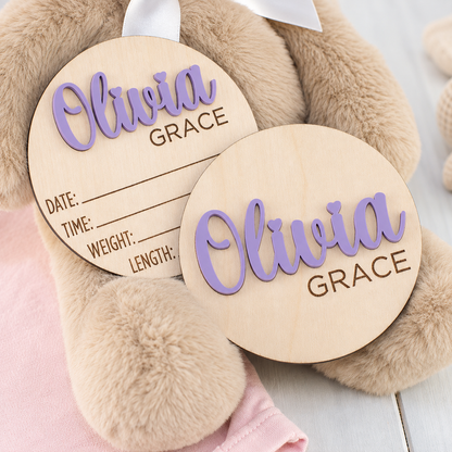 Two round wooden baby keepsake tags with personalized names 'Olivia' and 'Grace' in lavender script, displayed on a beige teddy bear with a pink blanket.