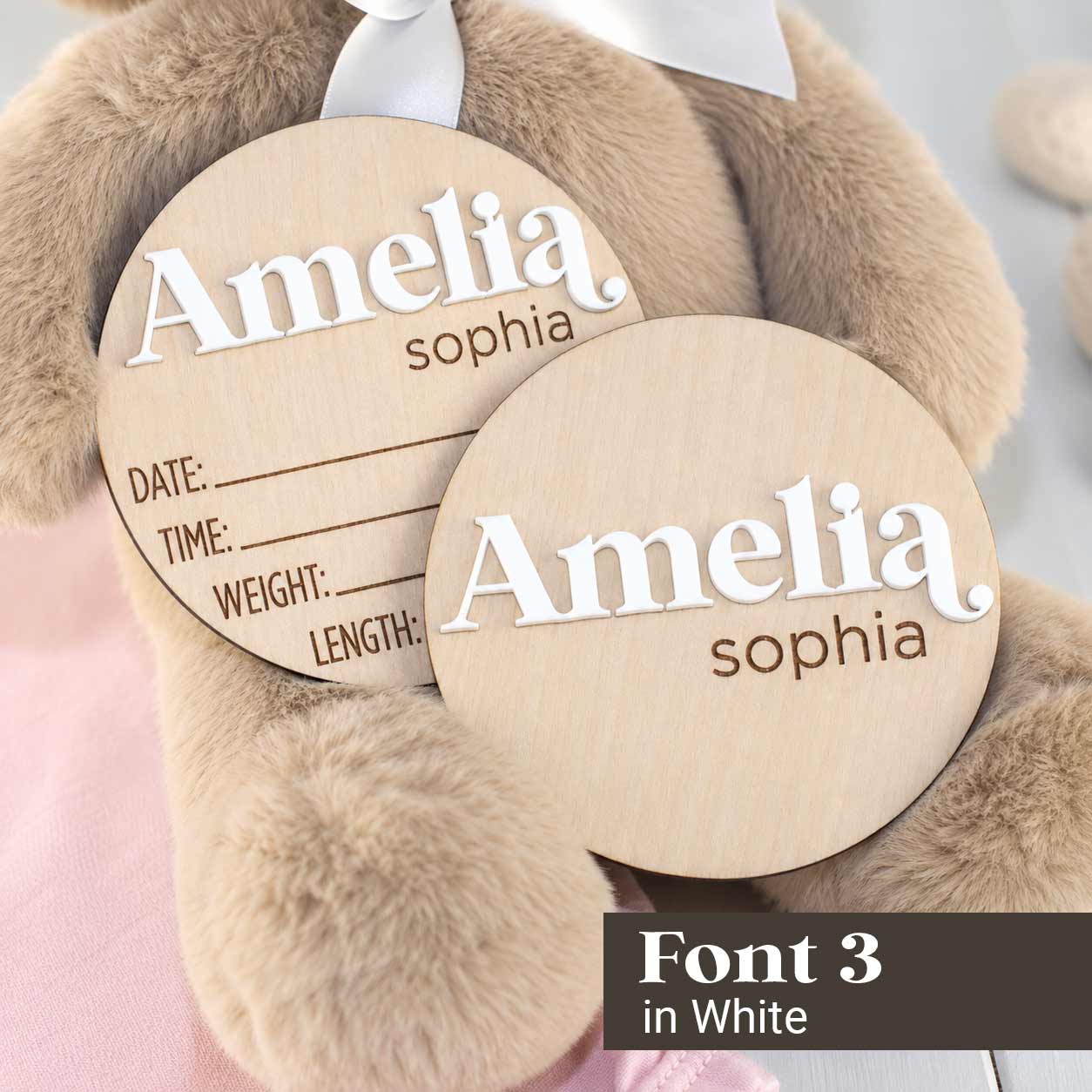 Two round wooden baby keepsake tags with engraved personalization reading 'Amelia sophia' and fields for date, time, weight, and length, shown on a plush teddy bear. Text at bottom right says 'Font 3 in White'.