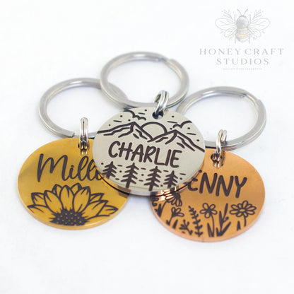 Collection of personalized stainless steel pet ID tags featuring mountain, sunflower, and wildflower designs in silver, gold, and copper finishes.