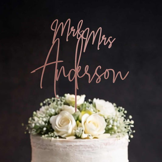 Rose gold mirror acrylic “Mr & Mrs Anderson” wedding cake topper on white floral wedding cake against dark background. Made by Honey Craft Studios