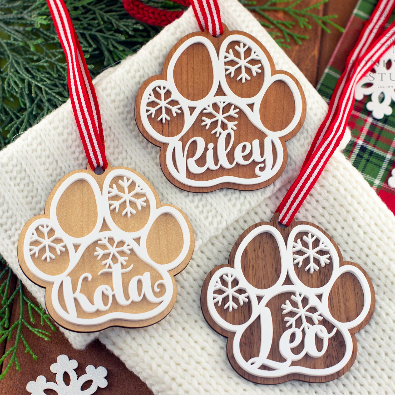 Three personalized wooden dog paw ornaments displayed on a cozy white stocking with red striped ribbons. Each ornament features a pet name — Riley, Kota, and Leo — with snowflake cutouts, showcasing the maple, cherry, and walnut wood options.