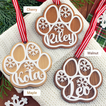 Labeled display of three personalized wooden dog paw ornaments showing the different wood types: maple, cherry, and walnut. Each ornament features a white acrylic snowflake design and custom pet name with red striped ribbon for hanging.