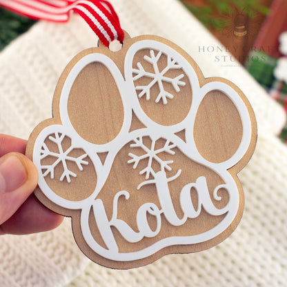 Close-up of a maple wood personalized dog paw ornament featuring the name “Kota” in white acrylic with snowflake accents and a red striped ribbon for hanging.