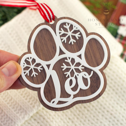 Close-up of a walnut wood personalized dog paw ornament featuring the name “Leo” in white acrylic with snowflake details and a festive red striped ribbon.