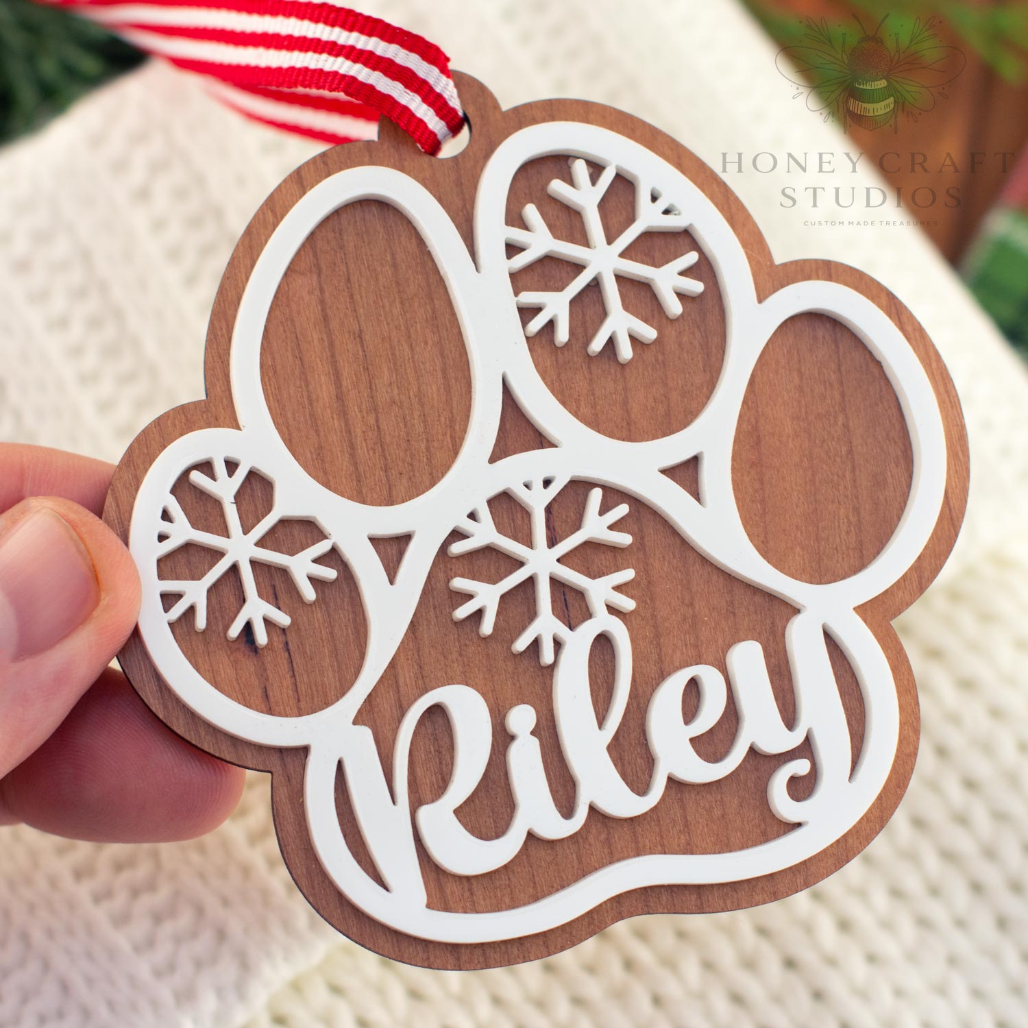 Close-up of a cherry wood personalized dog paw ornament featuring the name “Riley” in white acrylic lettering with snowflake cutouts and a red striped hanging ribbon.