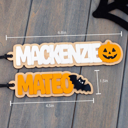 Two personalized wooden name tags with Halloween-themed lettering and accents, shown on a gray wood plank background with measurement lines and labels indicating dimensions.