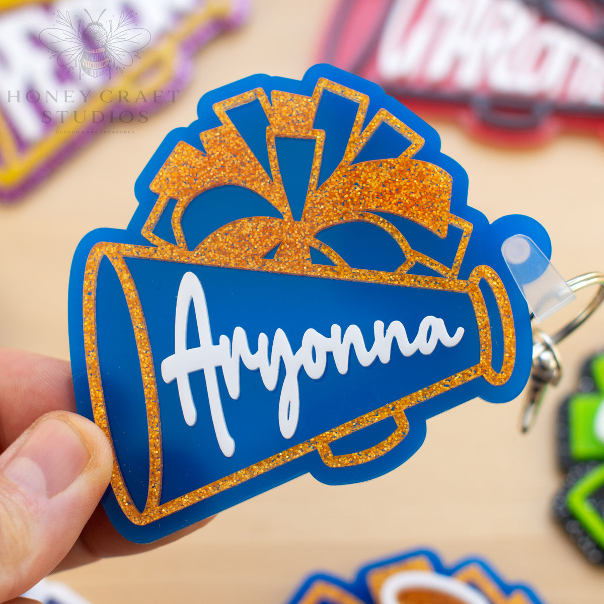 Blue and gold personalized cheer megaphone keychain featuring a custom name and glitter pom-pom design.