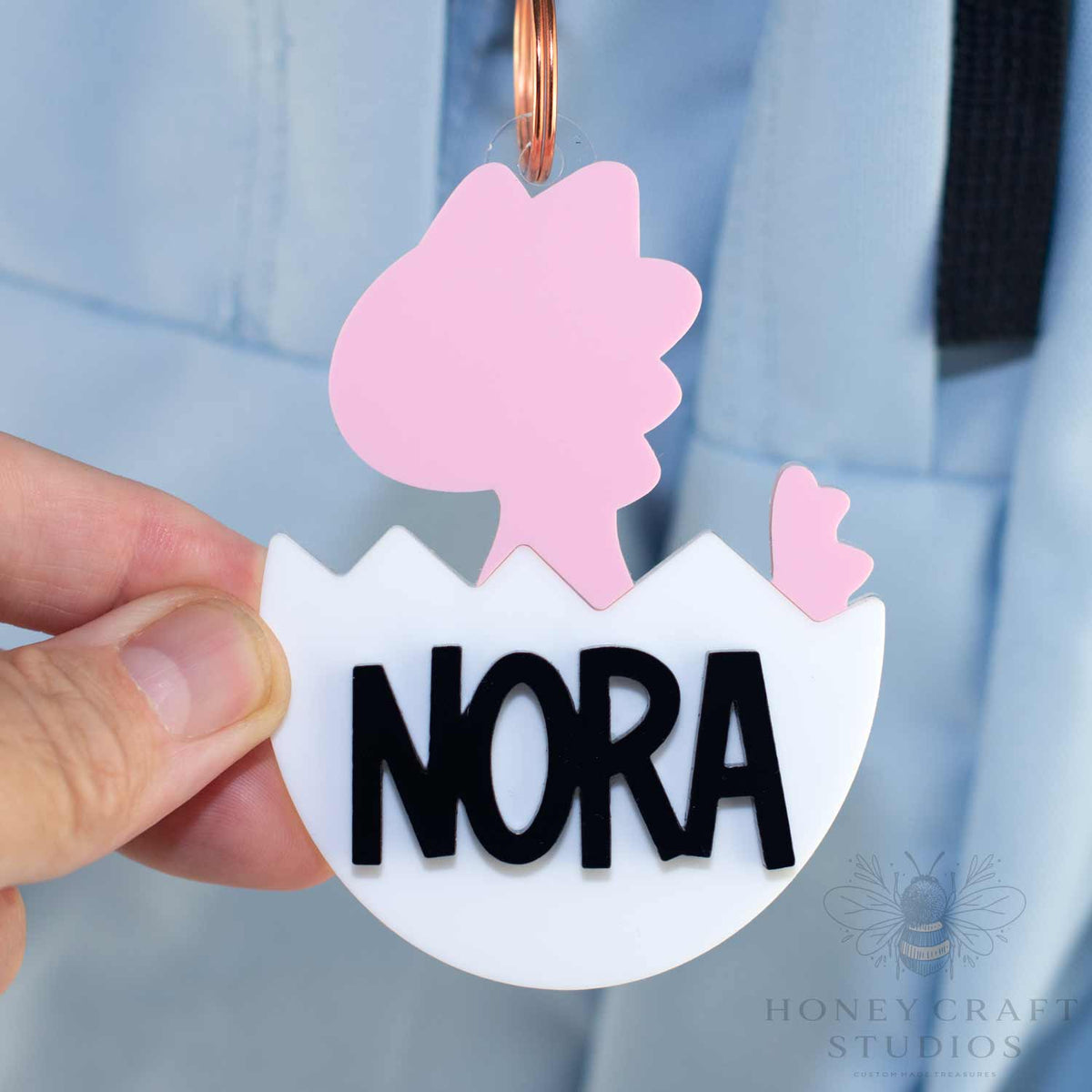 Pink baby dinosaur name tag with bold black custom name Nora in layered acrylic, hatching from a white egg.