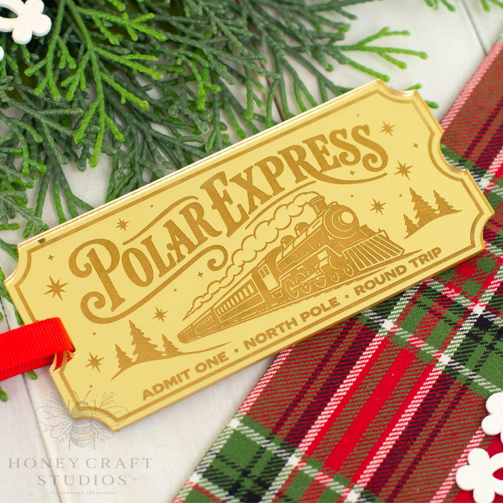 A decorative Polar Express train ticket tag with a red ribbon, styled on a white surface with green pine branches and a red plaid fabric. The tag features an engraved steam locomotive and holiday-themed illustrations.