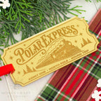 A decorative Polar Express train ticket tag with a red ribbon, styled on a white surface with green pine branches and a red plaid fabric. The tag features an engraved steam locomotive and holiday-themed illustrations.