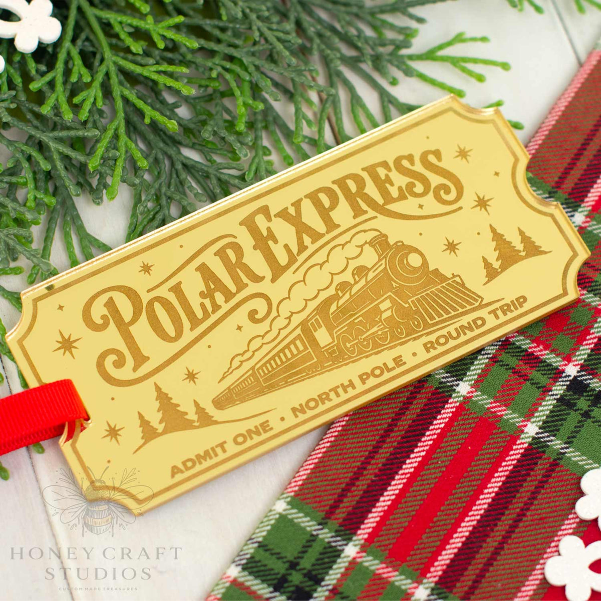A decorative Polar Express train ticket tag with a red ribbon, styled on a white surface with green pine branches and a red plaid fabric. The tag features an engraved steam locomotive and holiday-themed illustrations.