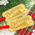 Two Polar Express train ticket ornaments by Honey Craft Studios, featuring a gold finish with engraved details and red ribbons, styled on a white surface with green pine branches and a red plaid blanket.