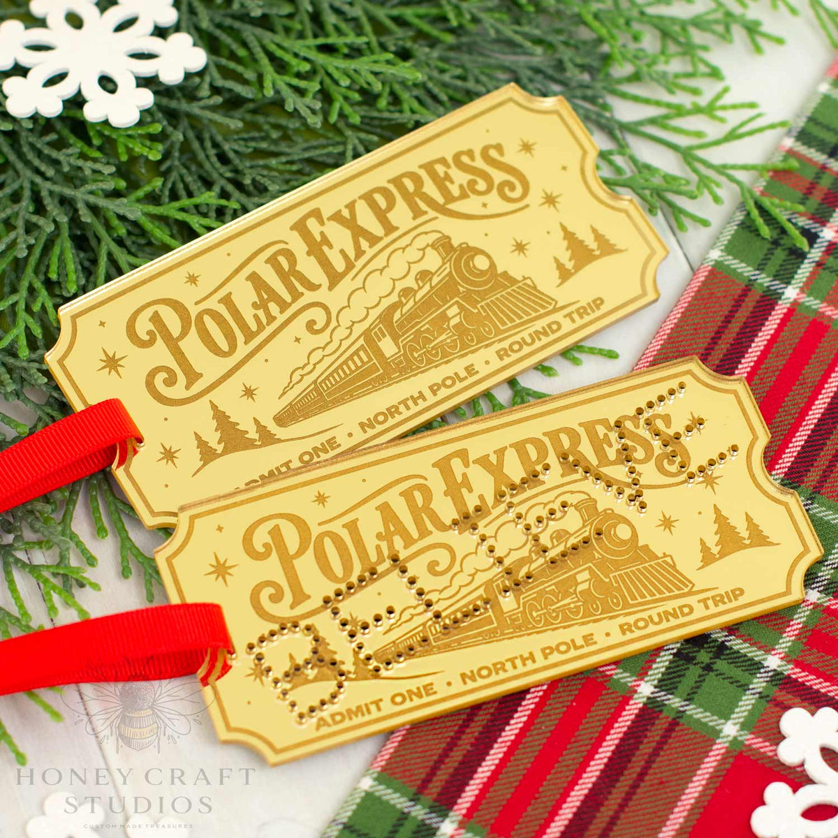 Two Polar Express train ticket ornaments by Honey Craft Studios, featuring a gold finish with engraved details and red ribbons, styled on a white surface with green pine branches and a red plaid blanket.