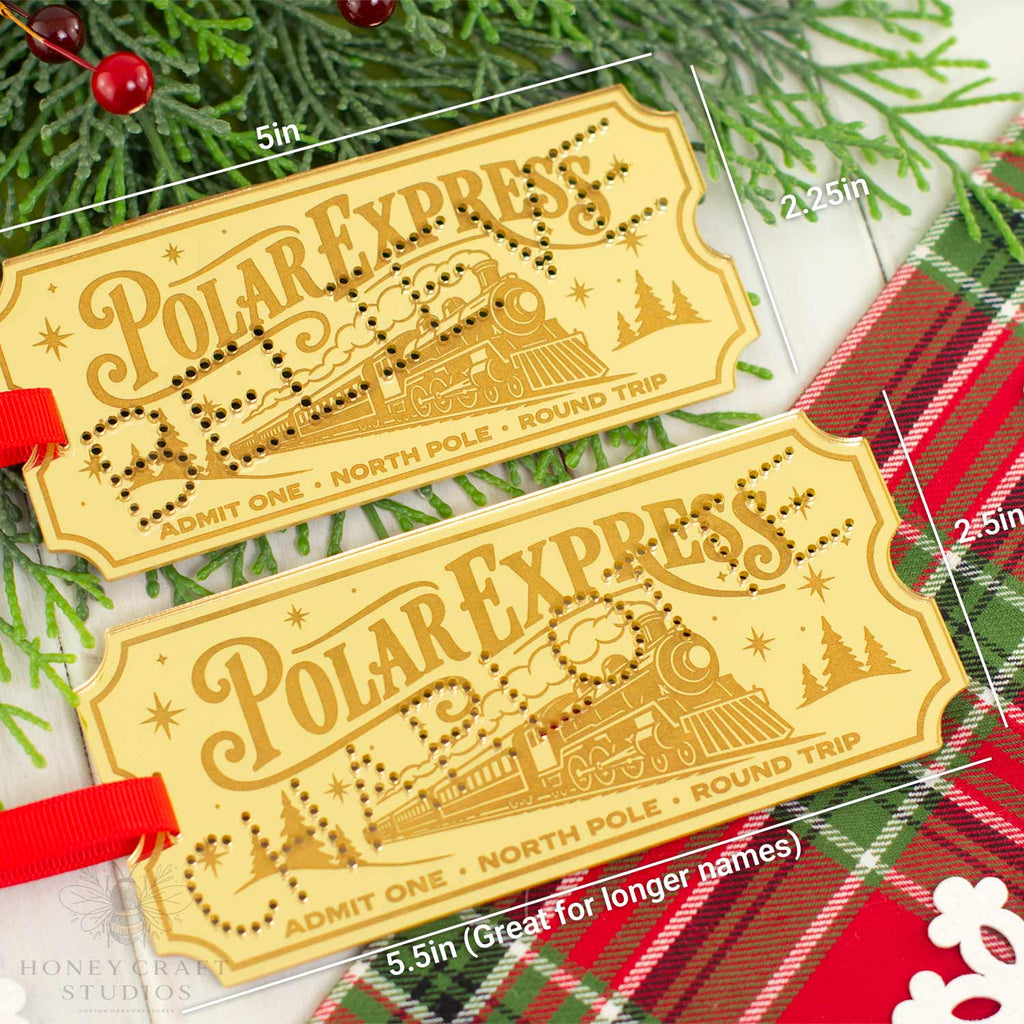 Two Polar Express train ticket luggage tags with red hanging ribbons, displayed on a festive background of pine branches and a red plaid blanket. White dimension lines and text indicate the size of the tags.