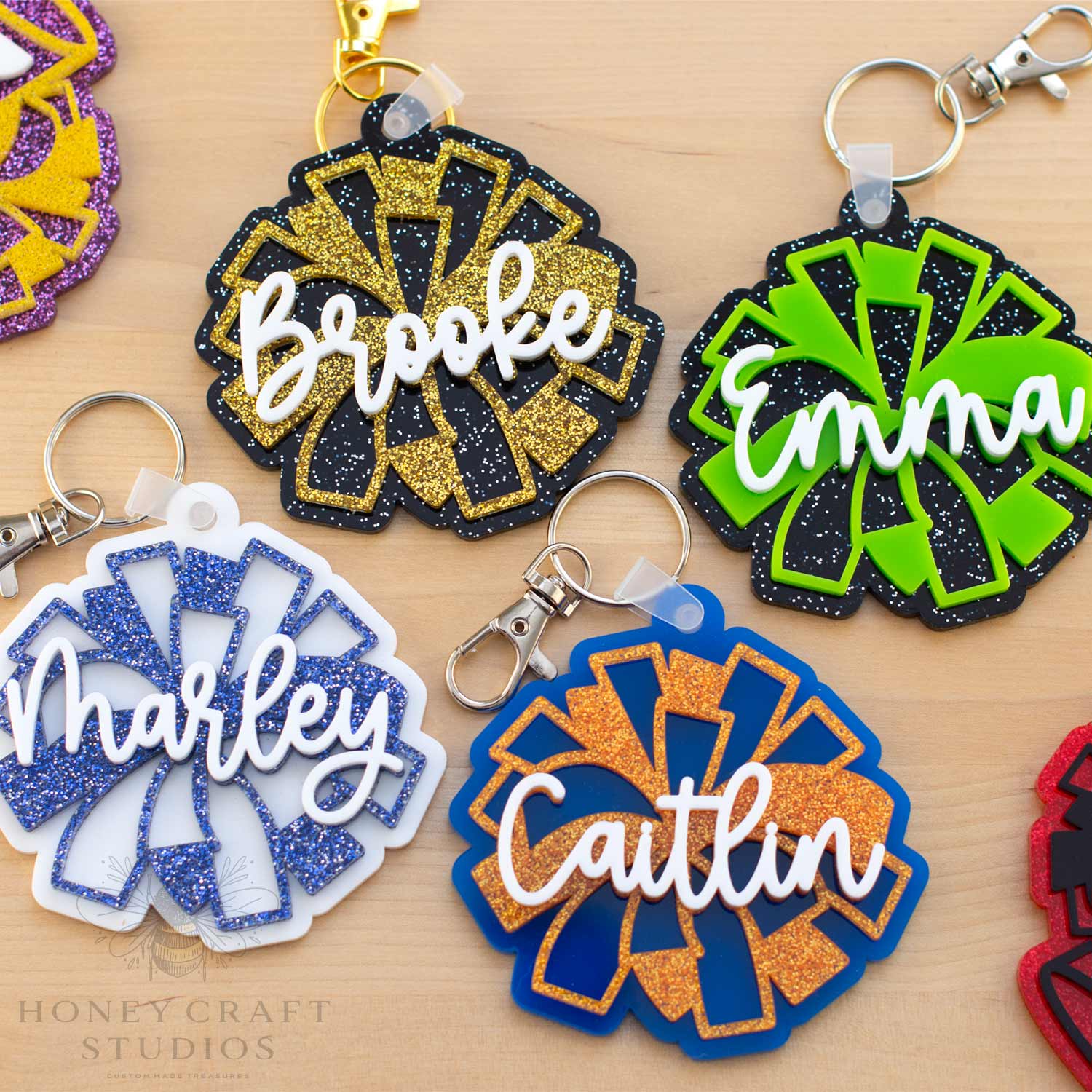 Collection of personalized cheer pom-pom name tag keychains in multiple team color combinations, including black and gold, lime green and black, blue and orange, and white and blue glitter acrylic designs.