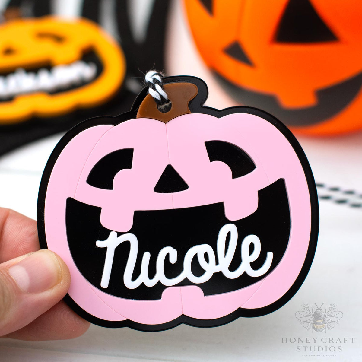 Pink pumpkin-shaped keychain with 'Nicole' text, held in front of pumpkins.