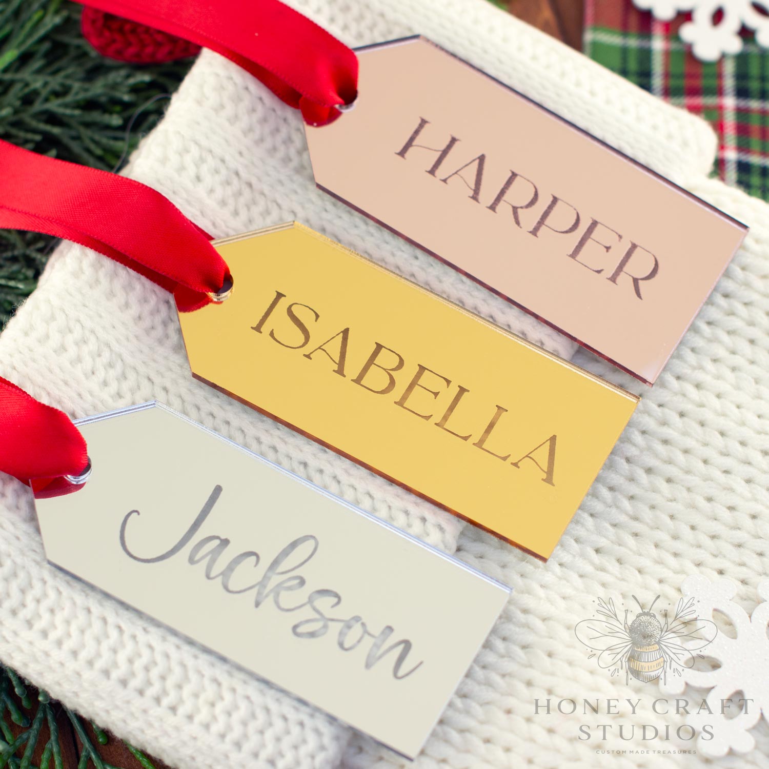 Set of three mirrored acrylic stocking name tags in silver, gold, and rose gold engraved with the names Jackson, Isabella, and Harper, tied with red ribbons.